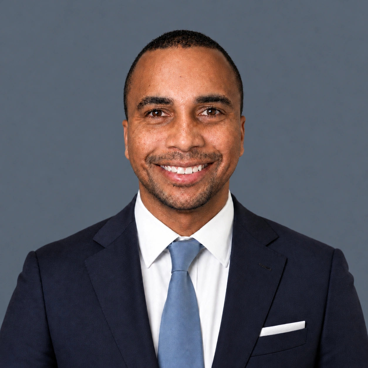Nate Smith in a navy suit, light blue tie, and white pocket square against a gray background